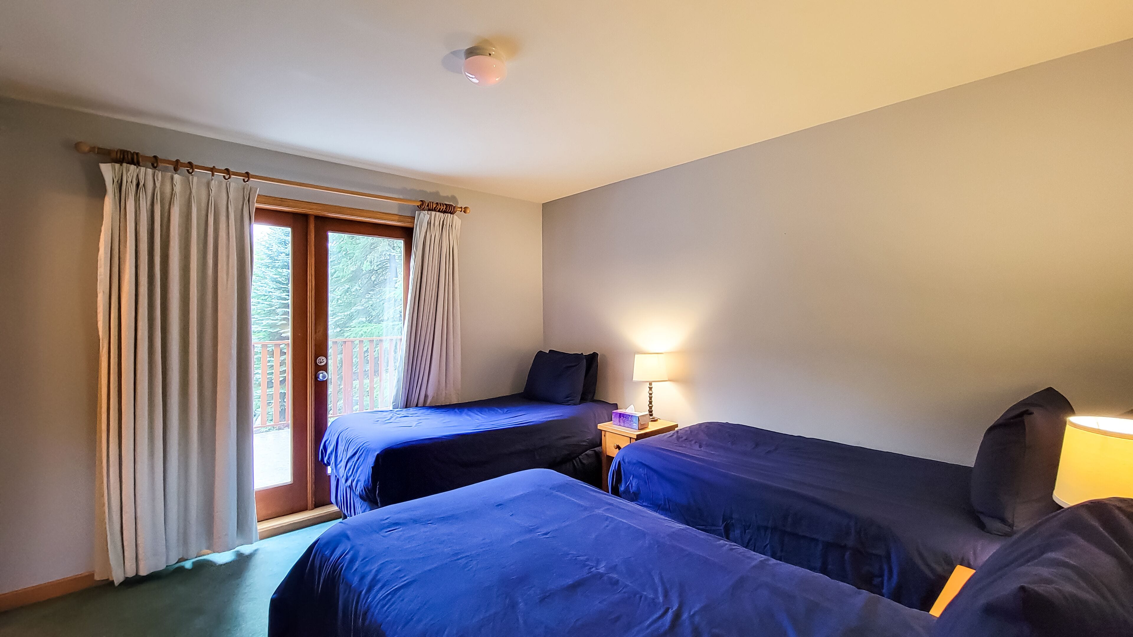 Une chambre avec deux lits, une fenêtre avec des rideaux, une table de chevet avec une lampe, et une vue sur les arbres à l’extérieur.