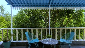 Un espace extérieur aménagé avec une zone de repos couverte, comprenant deux chaises et une table, le tout entouré d’une rambarde blanche et d’une végétation luxuriante.