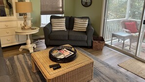 Un salon avec un canapé gris, des coussins à rayures, une table basse en osier sur laquelle sont posés des magazines et des télécommandes, une commode blanche et une vue sur l’extérieur grâce à une porte-fenêtre coulissante.