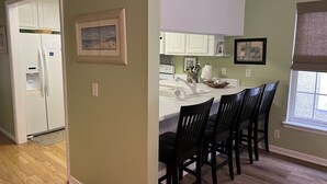Une cuisine avec un plan de travail blanc, des chaises noires et une fenêtre équipée de stores.