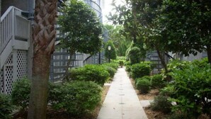 Un chemin bordé d’arbustes et d’arbres soigneusement taillés, qui mène à un bâtiment doté d’un escalier sur le côté gauche.