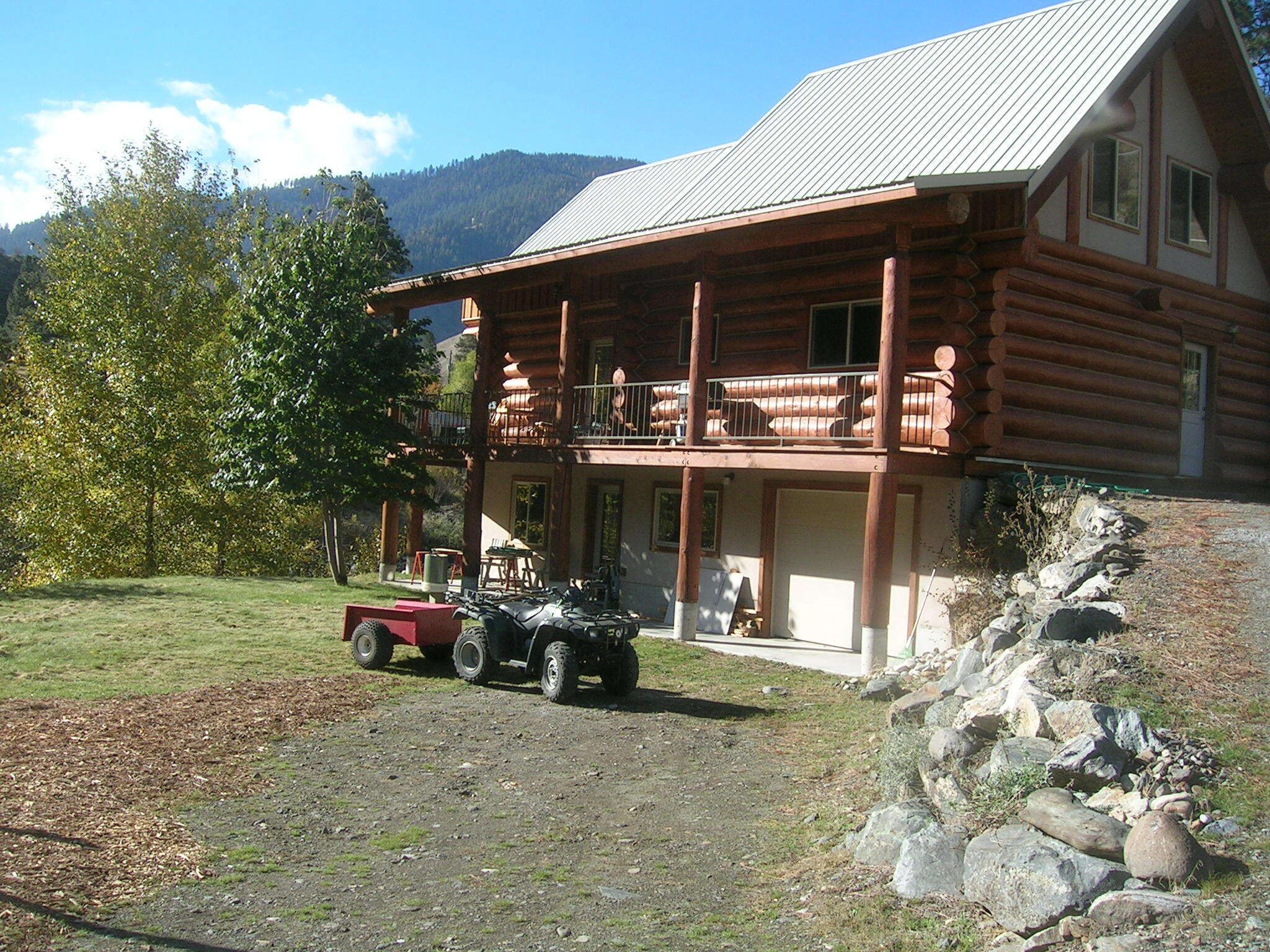 A log cabin with a balcony, a dirt driveway, and a mountainous backdrop.