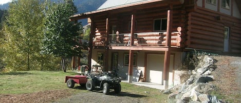 A log cabin with a balcony, a dirt driveway, and a mountainous backdrop.