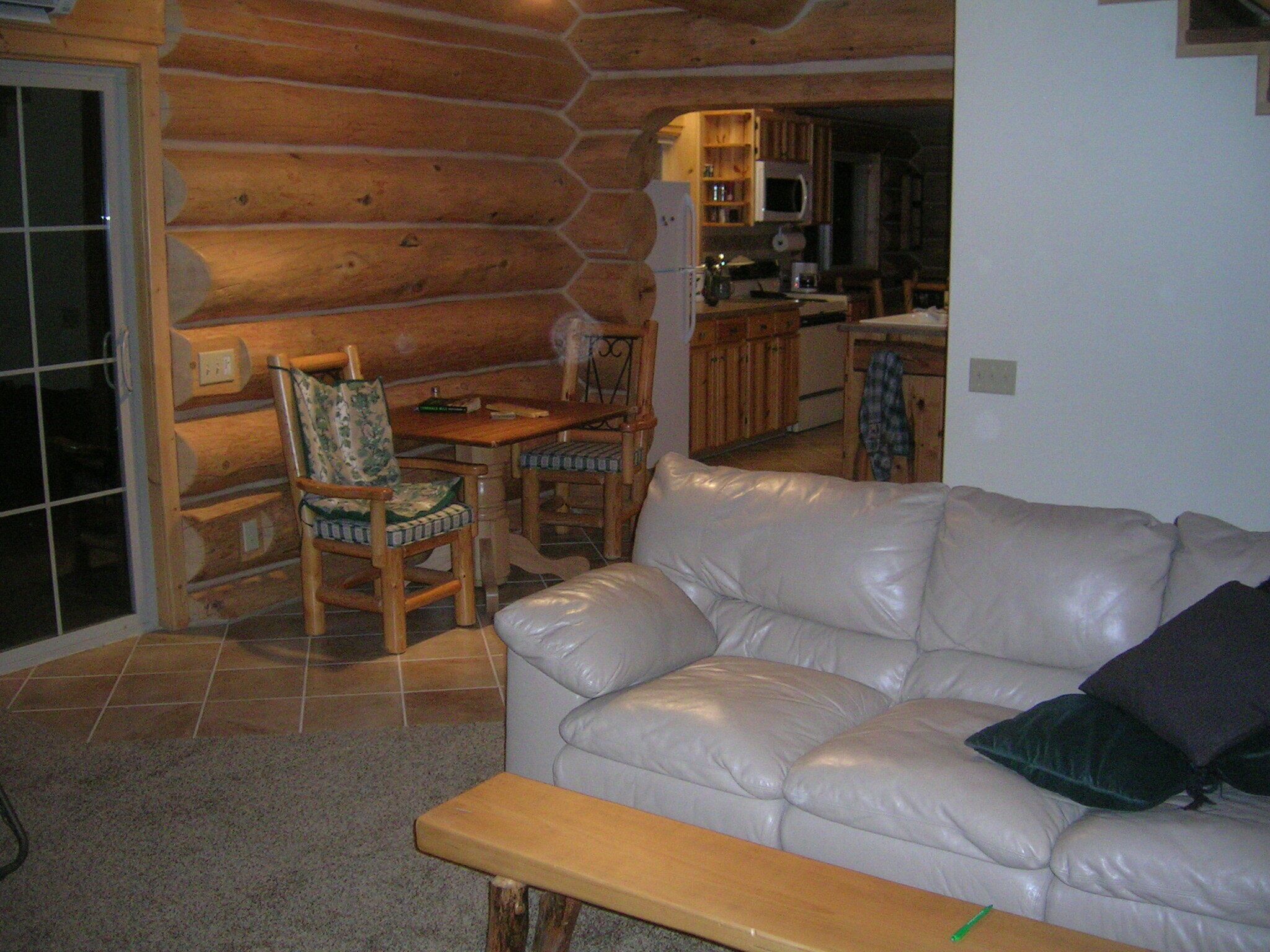 A log cabin interior with a white leather sofa, a wooden dining table with chairs, and a kitchen area visible through an archway.
