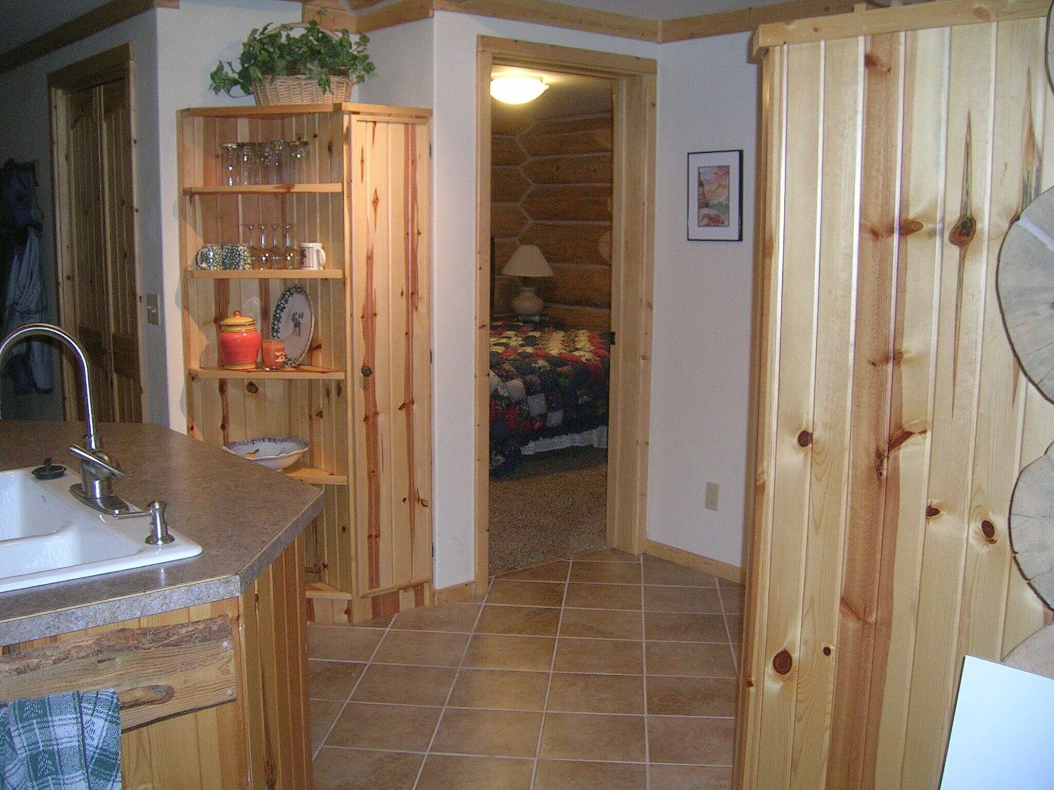Cuina amb armaris de fusta, un rentapeus i una encimera. Hi ha una porta que condueix a un dormitori amb un llit i una làmpada.