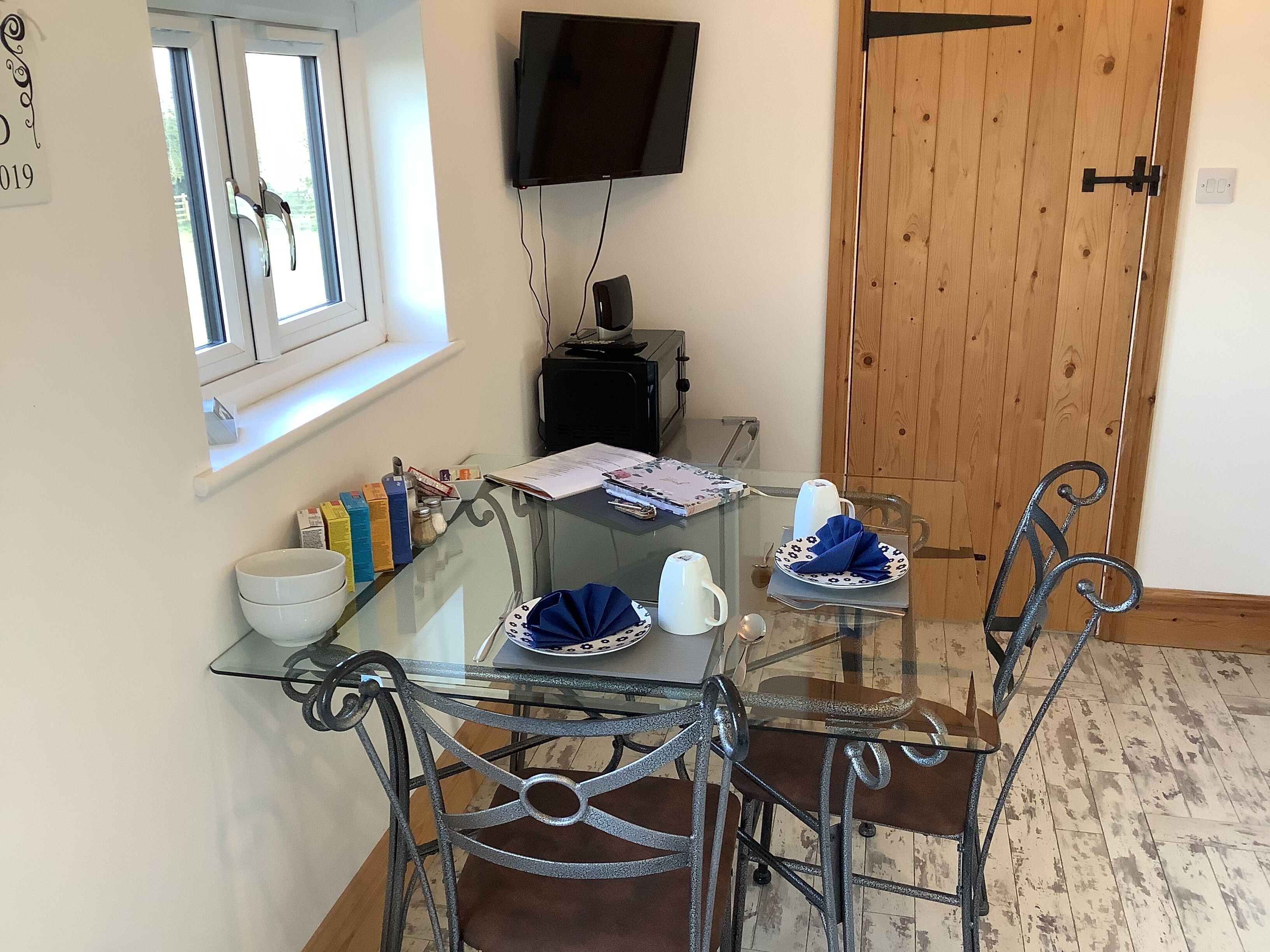 A dining area with a glass table, metal chairs, a wooden door, and a TV mounted on the wall.