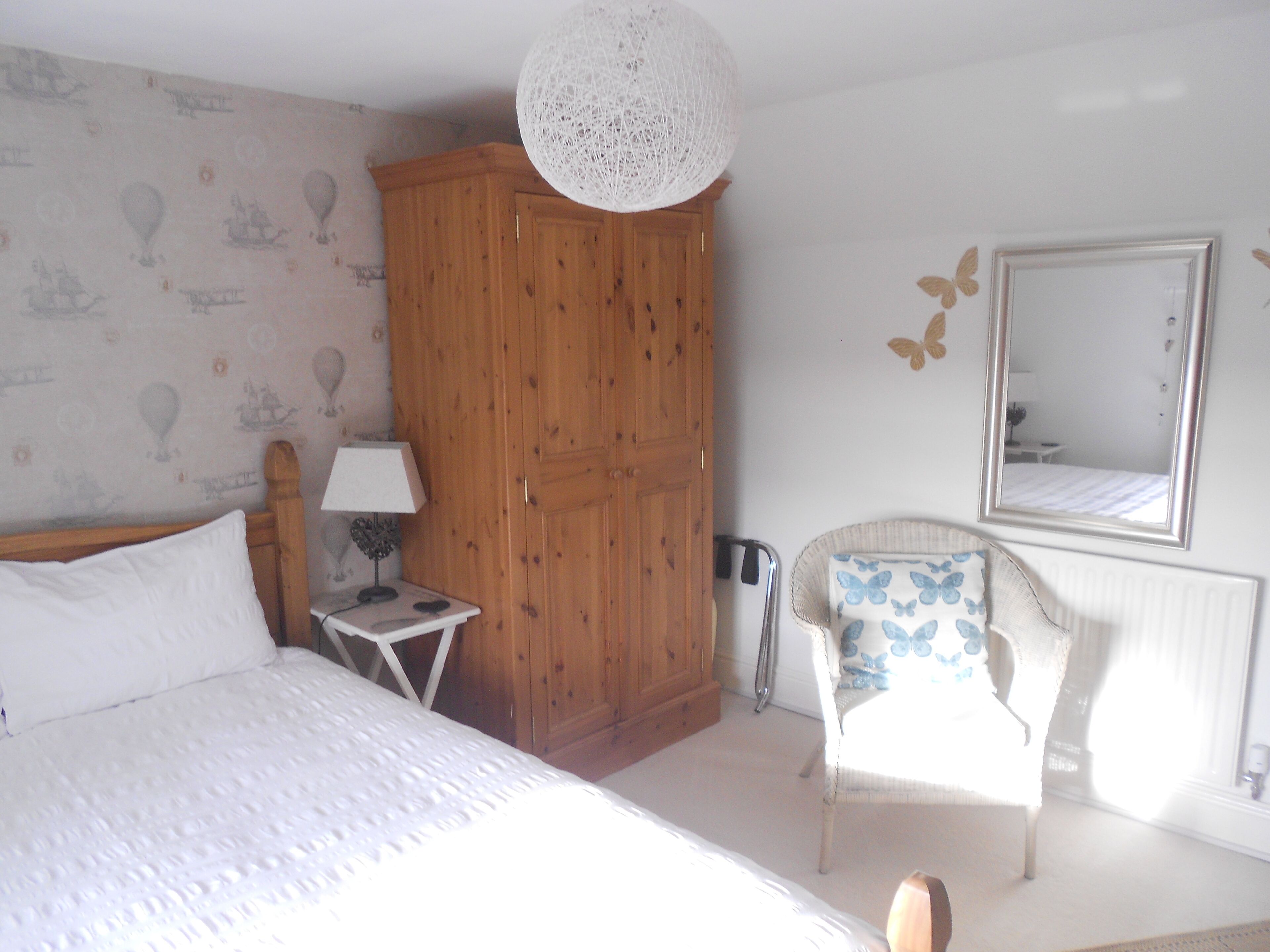 Een slaapkamer met een bed, een houten garderobe, een klein nachtkastje met een lamp, een stoel met een kussen in vlinder vorm, en een spiegel aan de muur.
