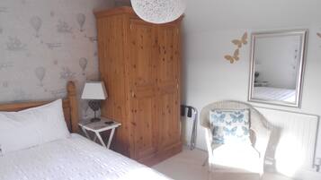 Een slaapkamer met een bed, een houten garderobe, een klein nachtkastje met een lamp, een stoel met een kussen in vlinder vorm, en een spiegel aan de muur.