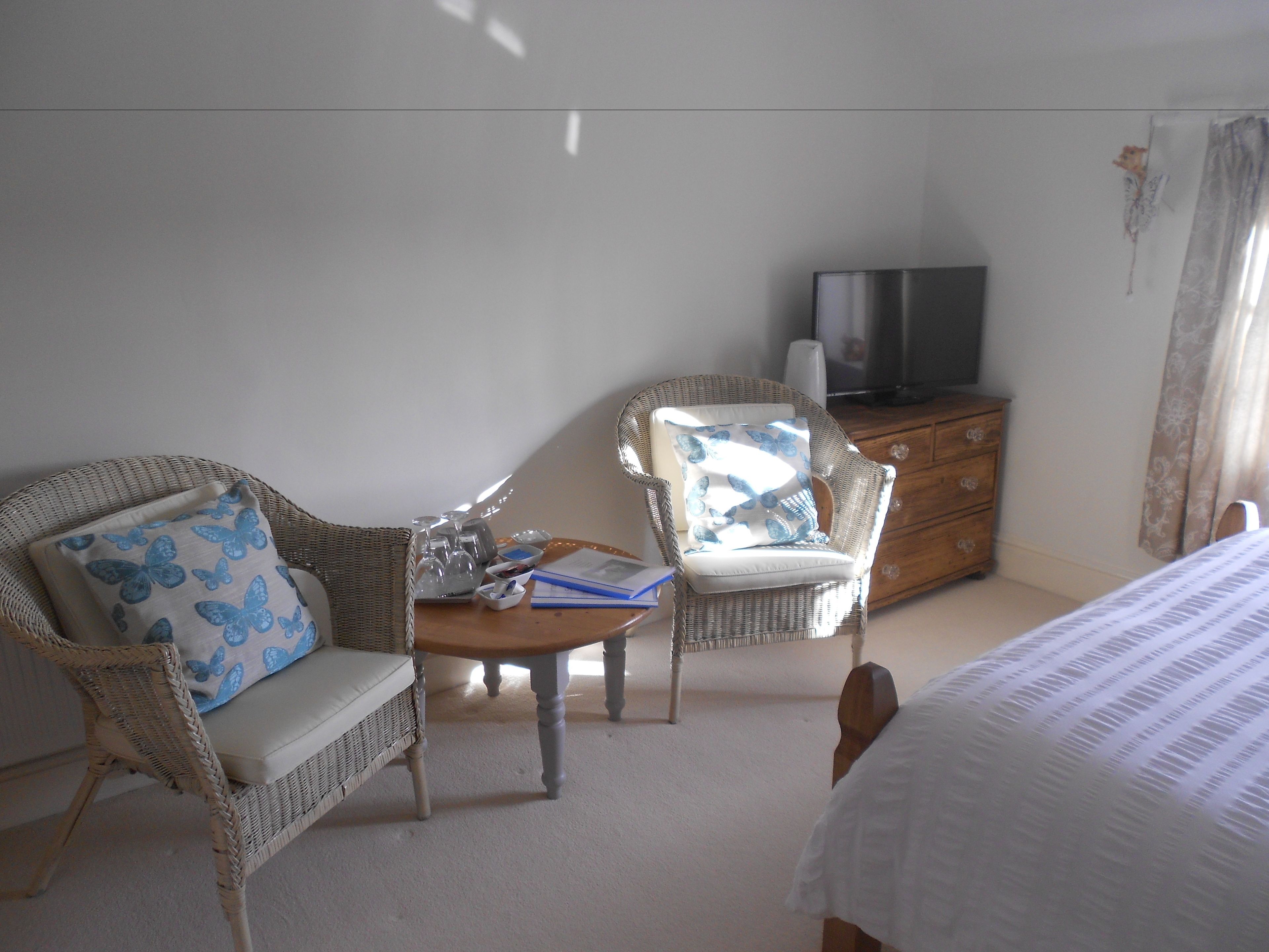 Un dormitorio con una cama, dos sillones de mimbre con cojines de diseño de mariposas, un aparador de madera, un televisor encima y una mesita con un recipiente de vidrio y documentos.