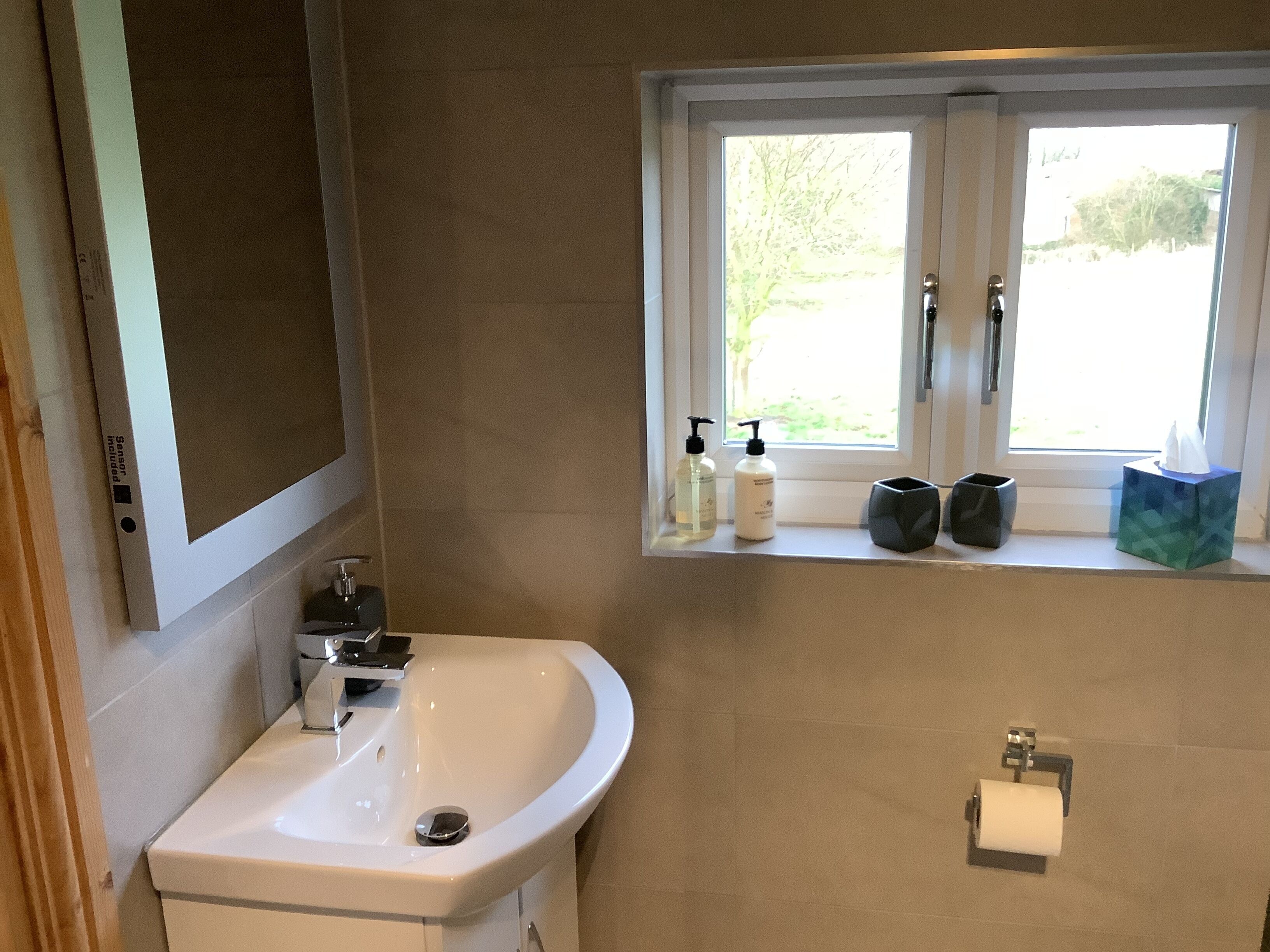 A bathroom with a white sink, a window with a view of greenery, and toiletries on the windowsill.