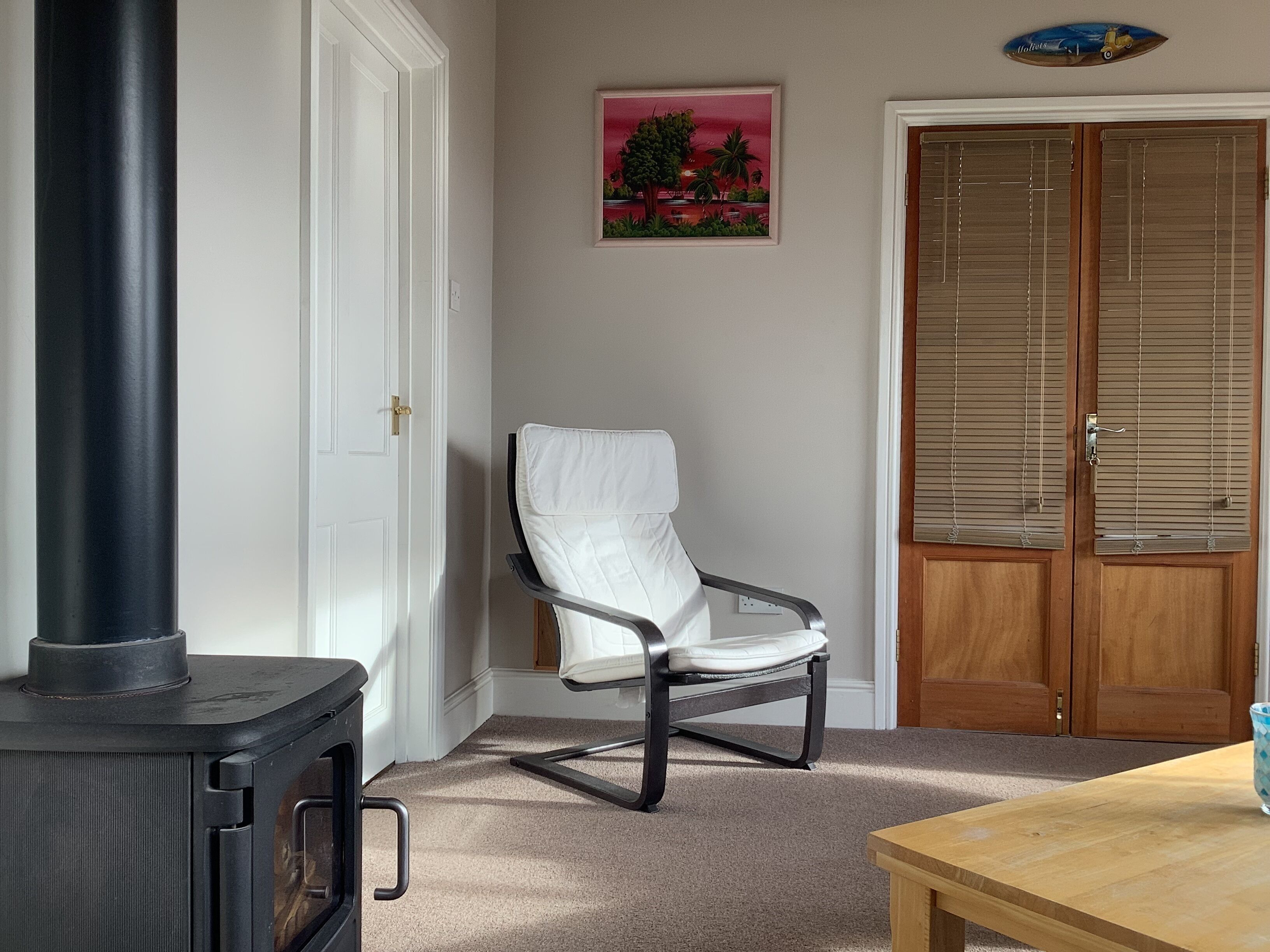 A room with a black wood-burning stove, a white upholstered chair, a wooden table, a wooden door with blinds, and a framed painting on the wall.