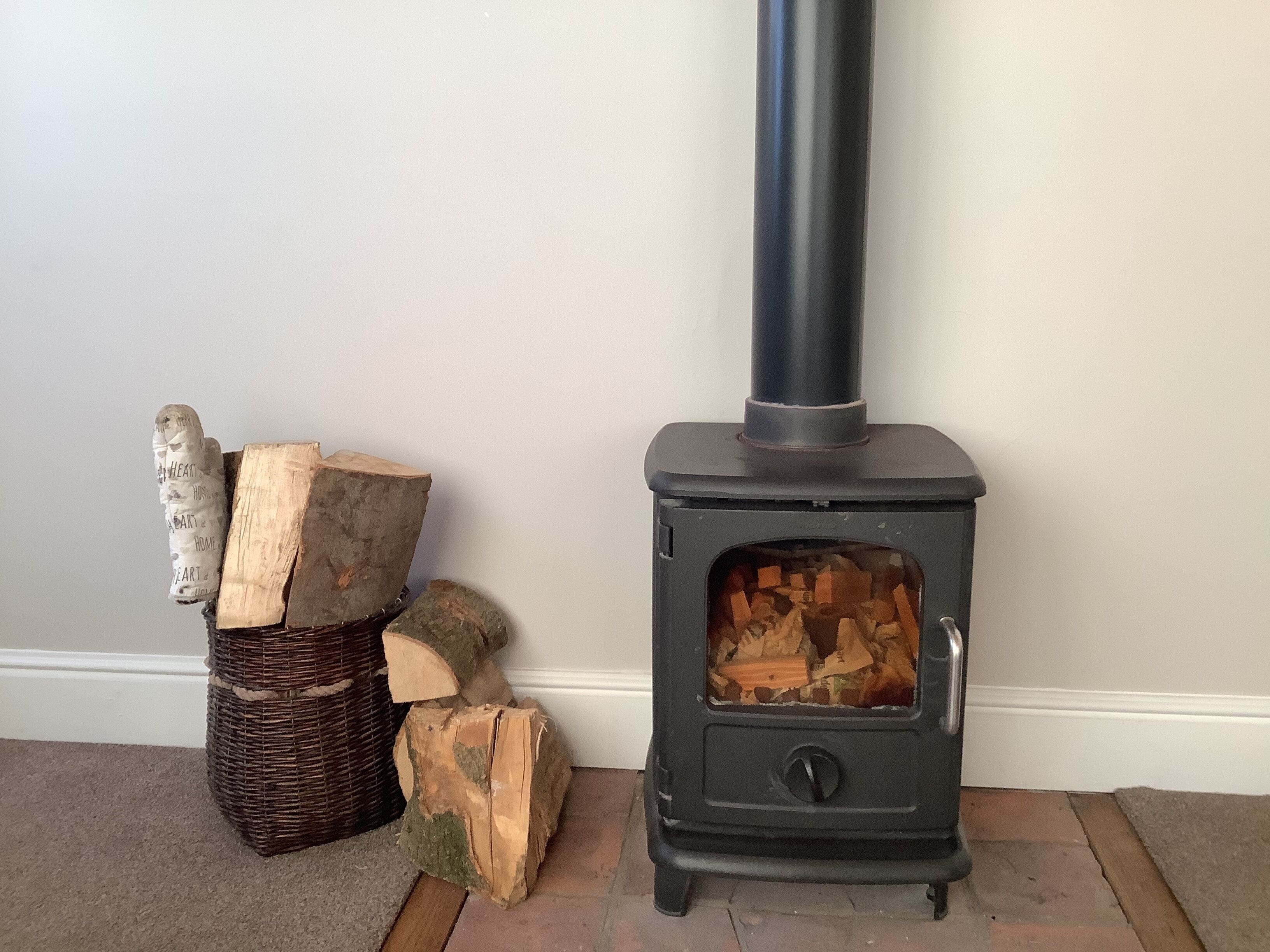 A wood-burning stove with logs inside, a wicker basket with logs, and a white wall.