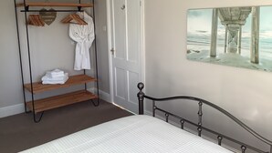 A bedroom with a bed, a wooden shelf with towels, a robe, and a painting of a pier. - Villa Farm Cottage (Ulceby)