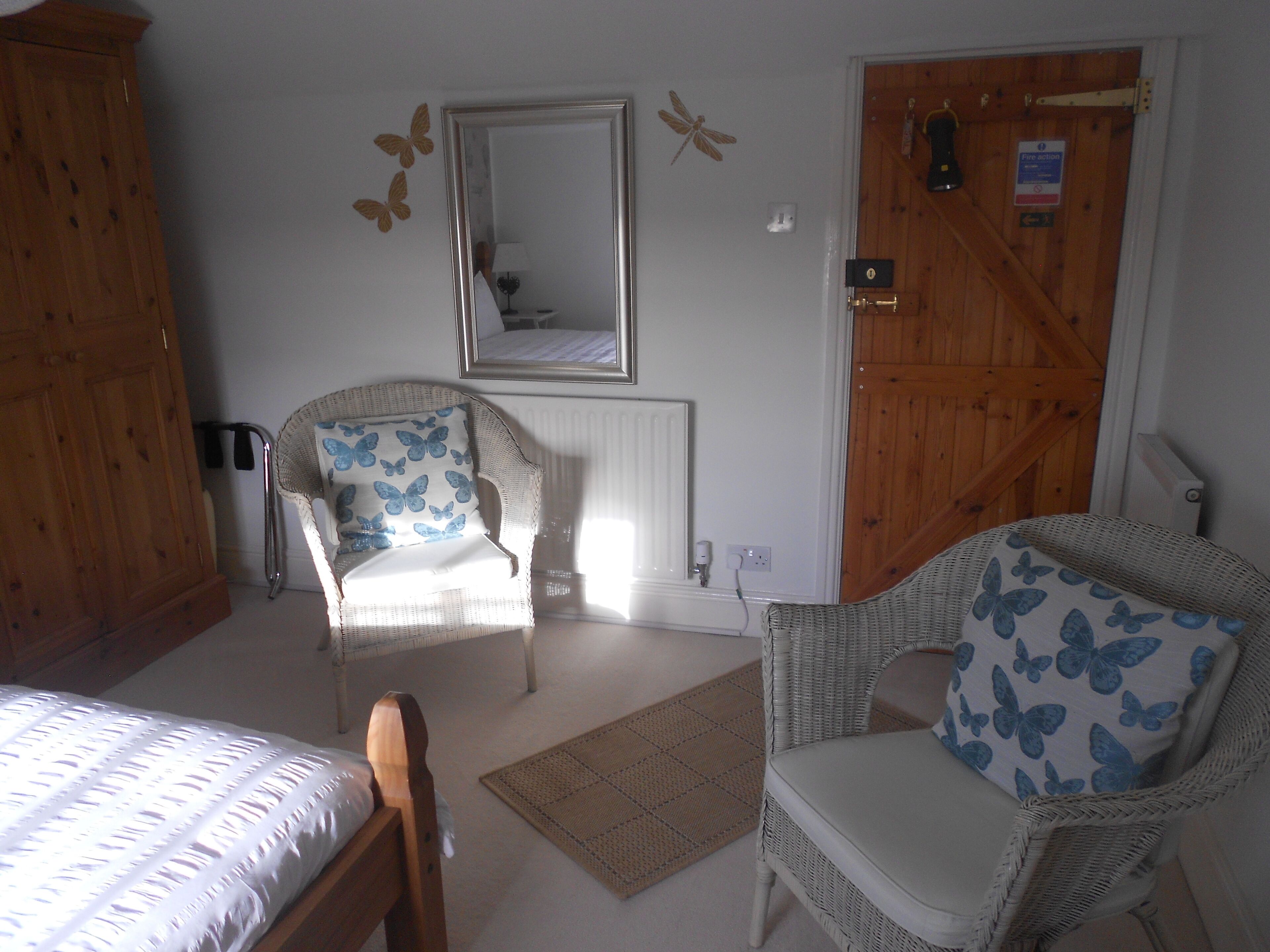A bedroom with two wicker chairs, a bed, a mirror, and a wooden door.