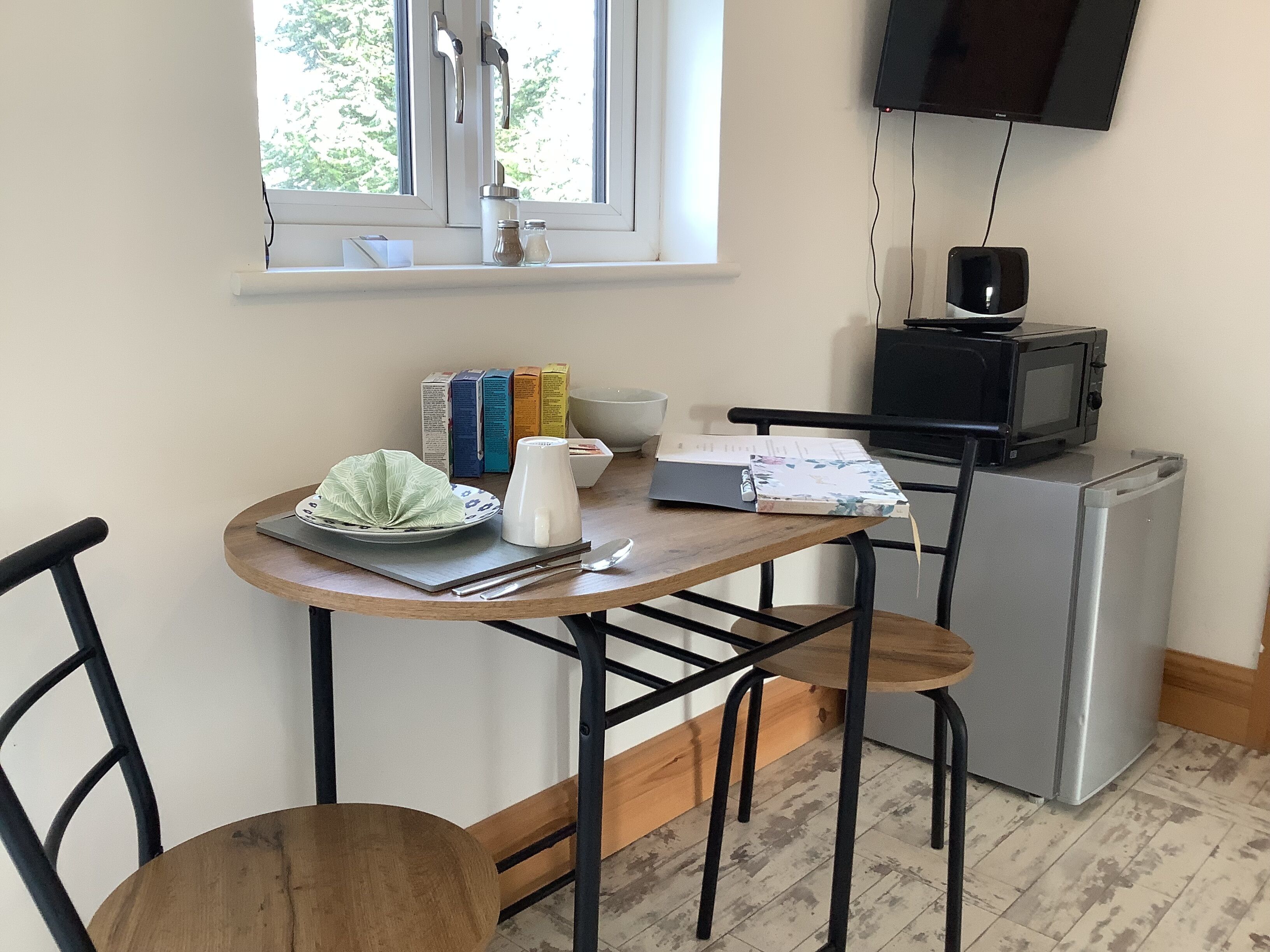 Un pequeño rincón de cocina con microondas, un refrigerador, una ventana con vistas a árboles, una mesa con un bol, un plato, una taza y algunos libros.