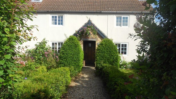 A traditional white building with a gravel pathway leading to a wooden door, surrounded by lush greenery and flowering plants. - Villa Farm Cottage (Ulceby)