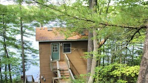 Une cabane en bois avec un escalier menant à une porte verte, entourée d’arbres et bordée par un plan d’eau.