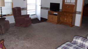 Een woonkamer met een bruine fauteuil, een televisie op een houten kast en een sofa met een geruit patroon.