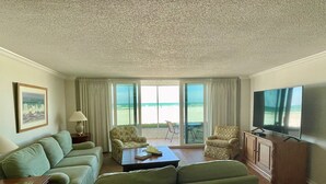 A living room with a sofa set, a coffee table, a television, and a view of the beach through large windows. - Crystal Sands Tower Unit 104 - Beachfront (Sarasota)