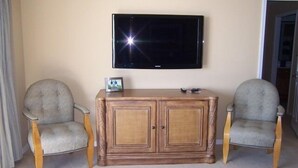 A living room with two armchairs, a wooden cabinet, and a flat-screen TV mounted on the wall. - Crystal Sands Tower Unit 104 - Beachfront (Sarasota)