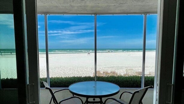A glass-enclosed patio with a round table and four chairs, overlooking a beach with a clear sky. - Crystal Sands Tower Unit 104 - Beachfront (Sarasota)