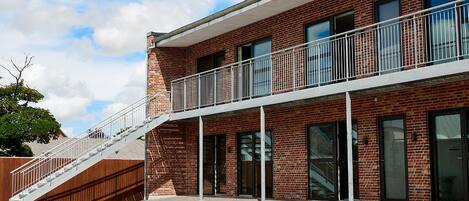 A modern building with a brick facade, a balcony with metal railings, and a staircase leading to the entrance.