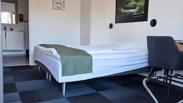 Habitación de hotel con cama, escritorio, silla, un cuadro en la pared y una cocina visible a través de una puerta abierta.