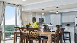 Un comedor con una mesa y sillas de madera, un jarrón de flores amarillas y vistas al exterior a través de amplias ventanas.