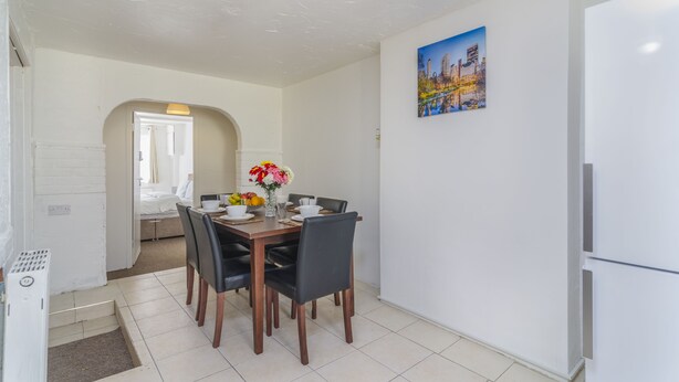 Un coin repas avec une table en bois, des chaises noires et un vase de fleurs. On y trouve également un réfrigérateur blanc et un tableau accroché au mur.