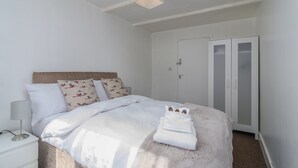 Un lit bien fait, avec du linge de lit blanc, un tapis blanc moelleux et une table de chevet blanche sur laquelle se trouve une lampe.