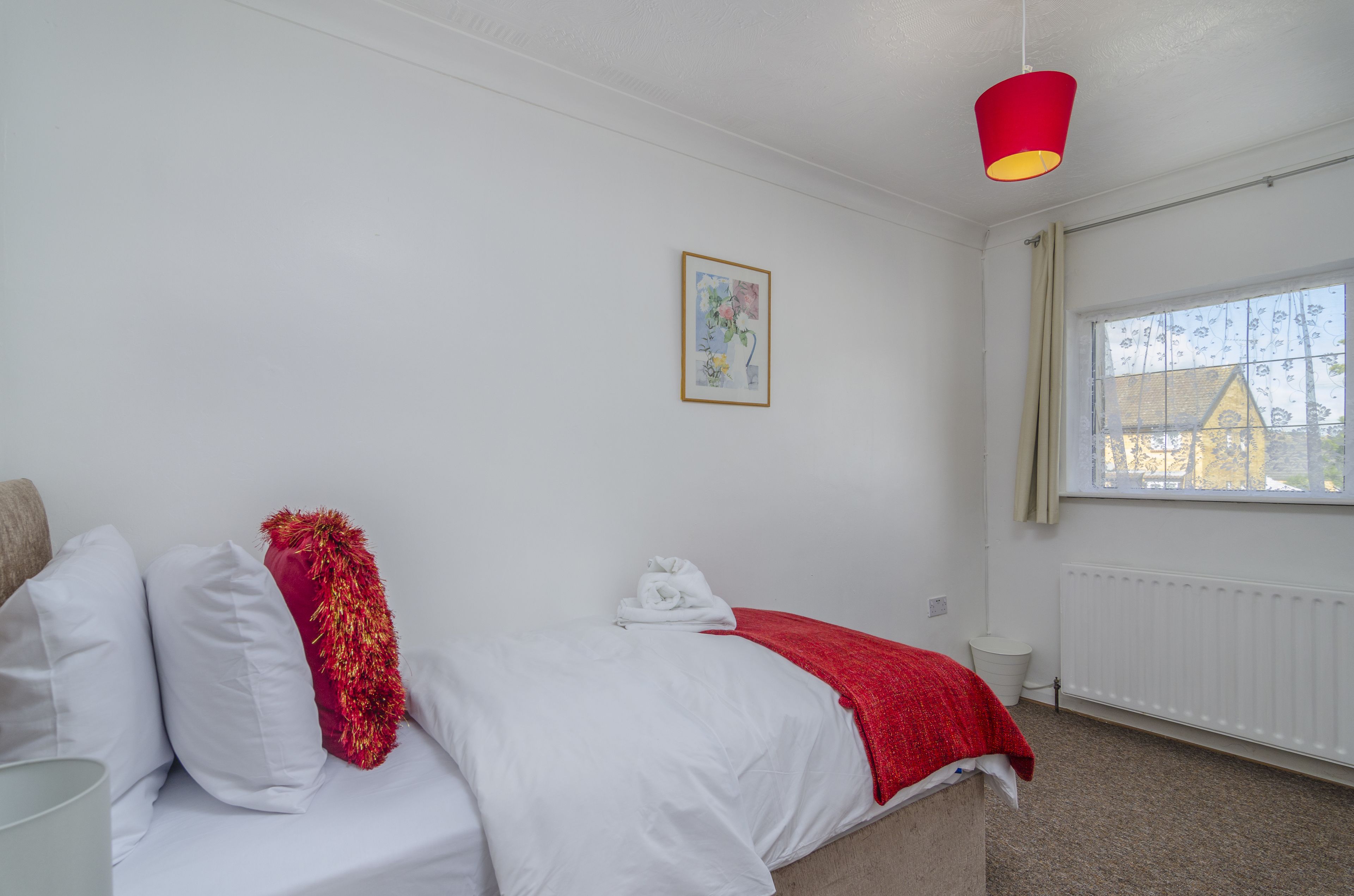 Una camera da letto con un letto, un cuscino decorativo rosso, un asciugamano bianco, una lampada a sospensione rossa, una finestra con tende e un quadro appeso al muro.