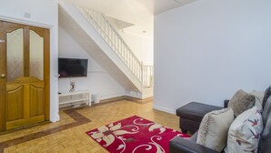 Un salon avec une porte en bois, un escalier, un téléviseur à écran plat, un tapis rouge à motifs et un canapé en cuir avec des coussins.