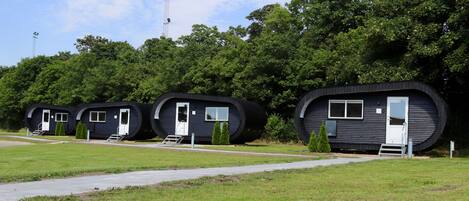 A row of cylindrical, dark-colored cabins with white doors and windows, surrounded by green grass and trees.