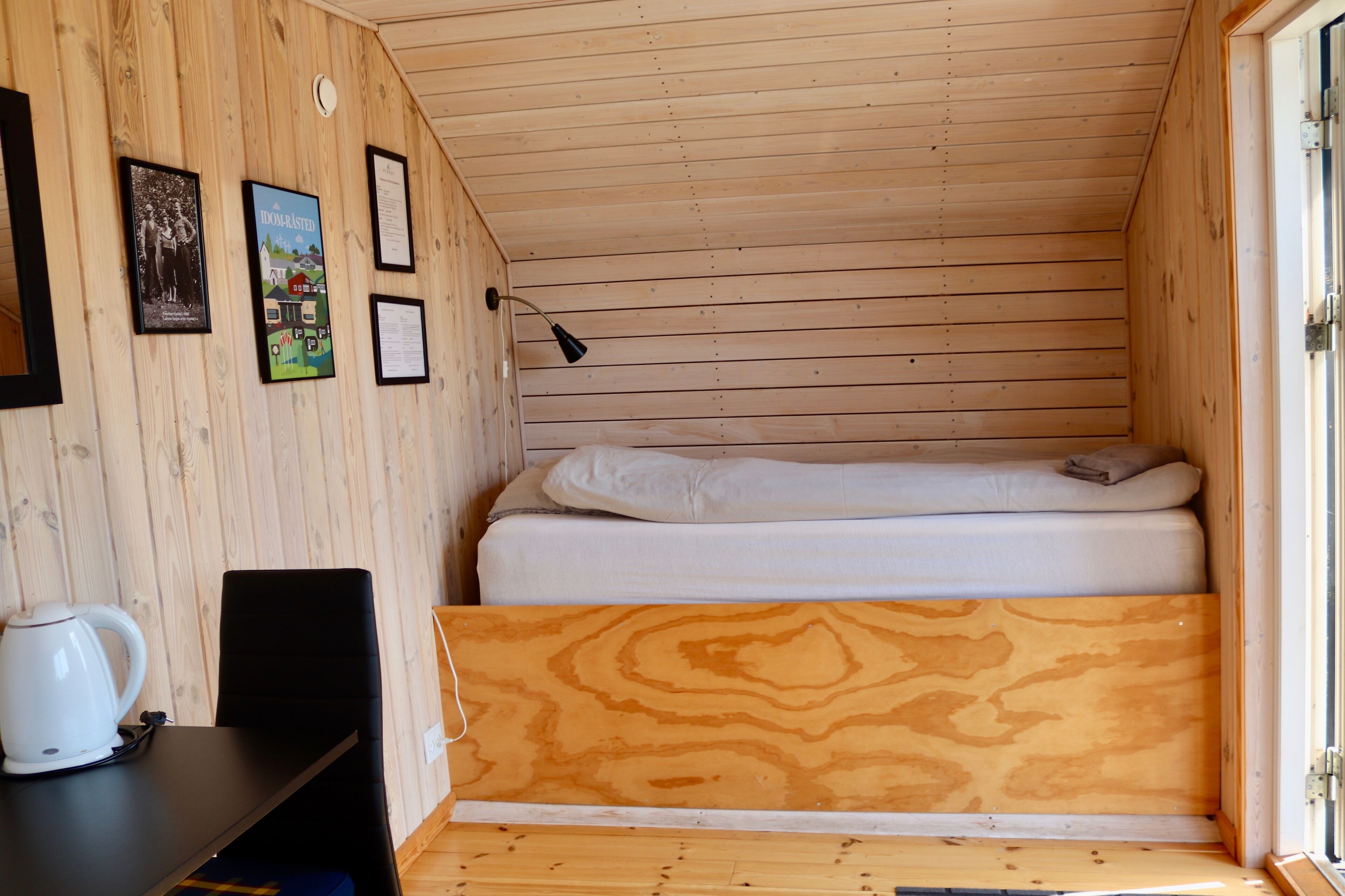 A small, wooden room with a bed, a desk, and a chair. There are framed pictures on the wall.