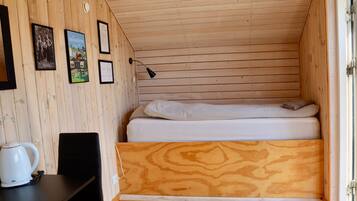 A small, wooden room with a bed, a desk, and a chair. There are framed pictures on the wall.