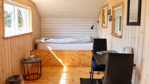 A compact room with a bed, a desk, and a chair. There is a window on one side and a door on the other. - Plexus Hytterne (Holstebro)