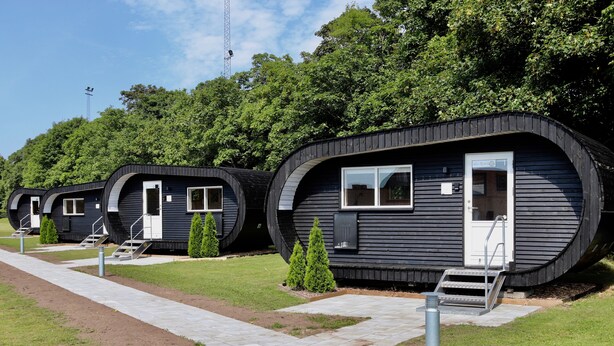A row of modern, curved cabins with external steps and small trees in front. - Plexus Hytterne (Holstebro)