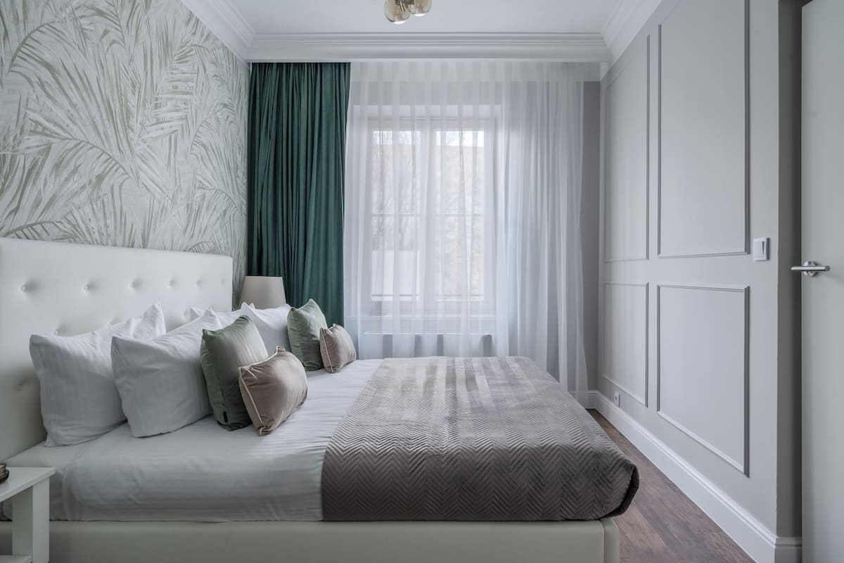 A neatly arranged bedroom with a large bed, a bedside table, and a window with sheer curtains.