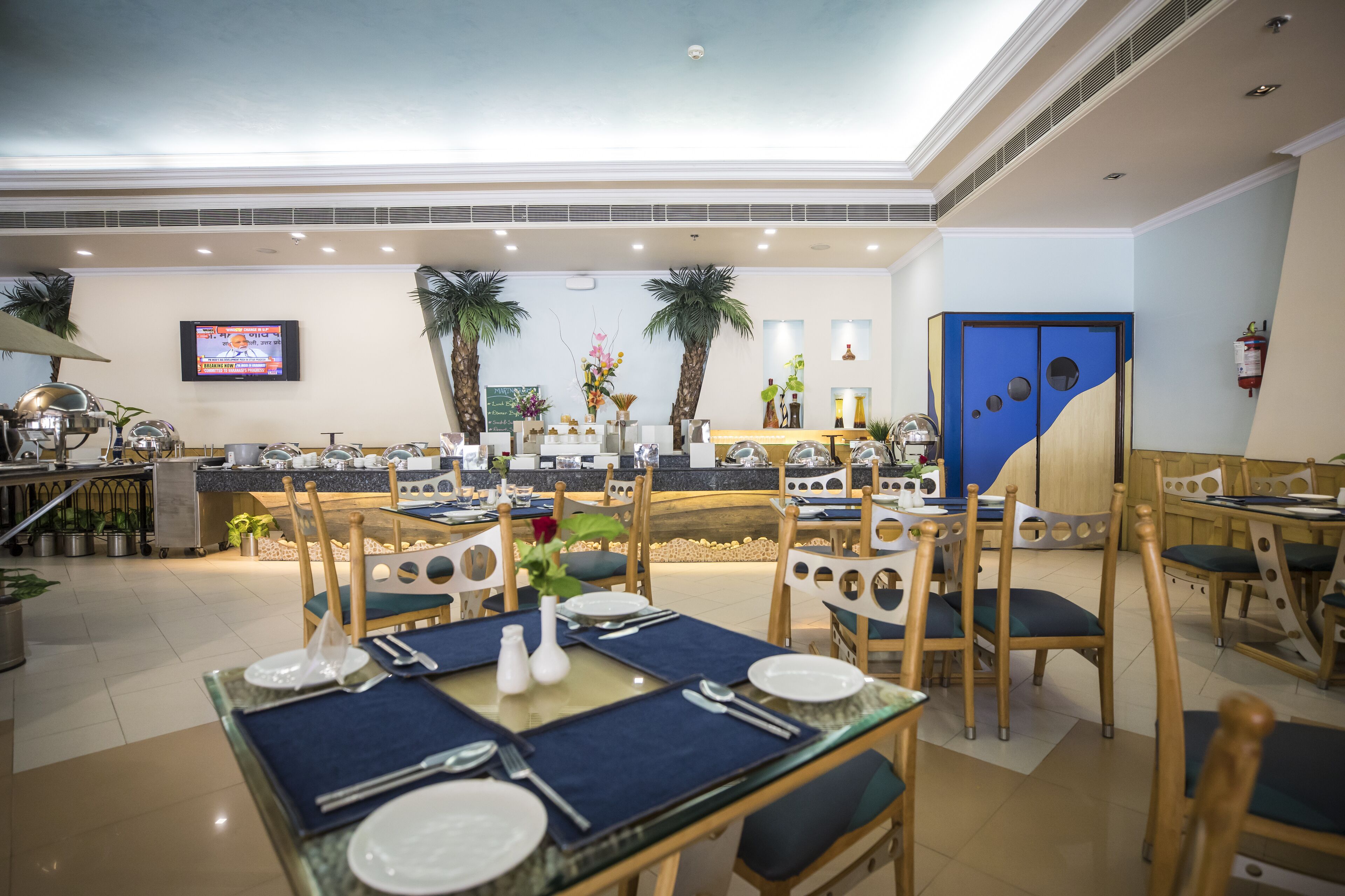 Un comedor de hotel con varias mesas dispuestas para comer, una pantalla de televisión y una zona de cocina al fondo.