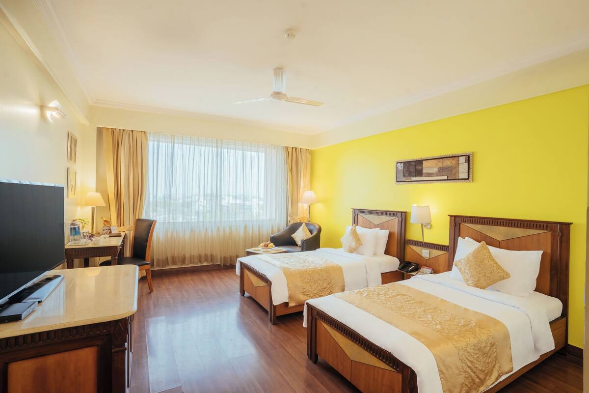 Quarto de hotel com duas camas, uma escrivaninha, uma cadeira, uma televisão e um ventilador de teto.