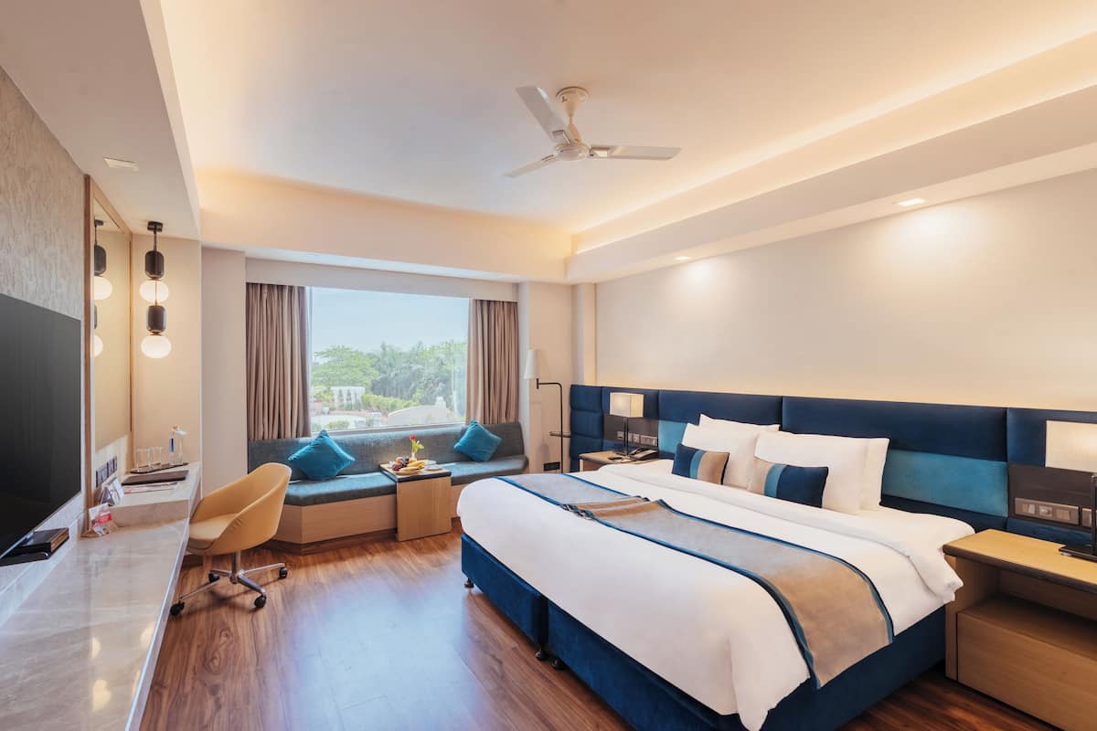 Quarto de hotel moderno com uma cama grande, escrivaninha, cadeira, televisão e janela com cortinas.