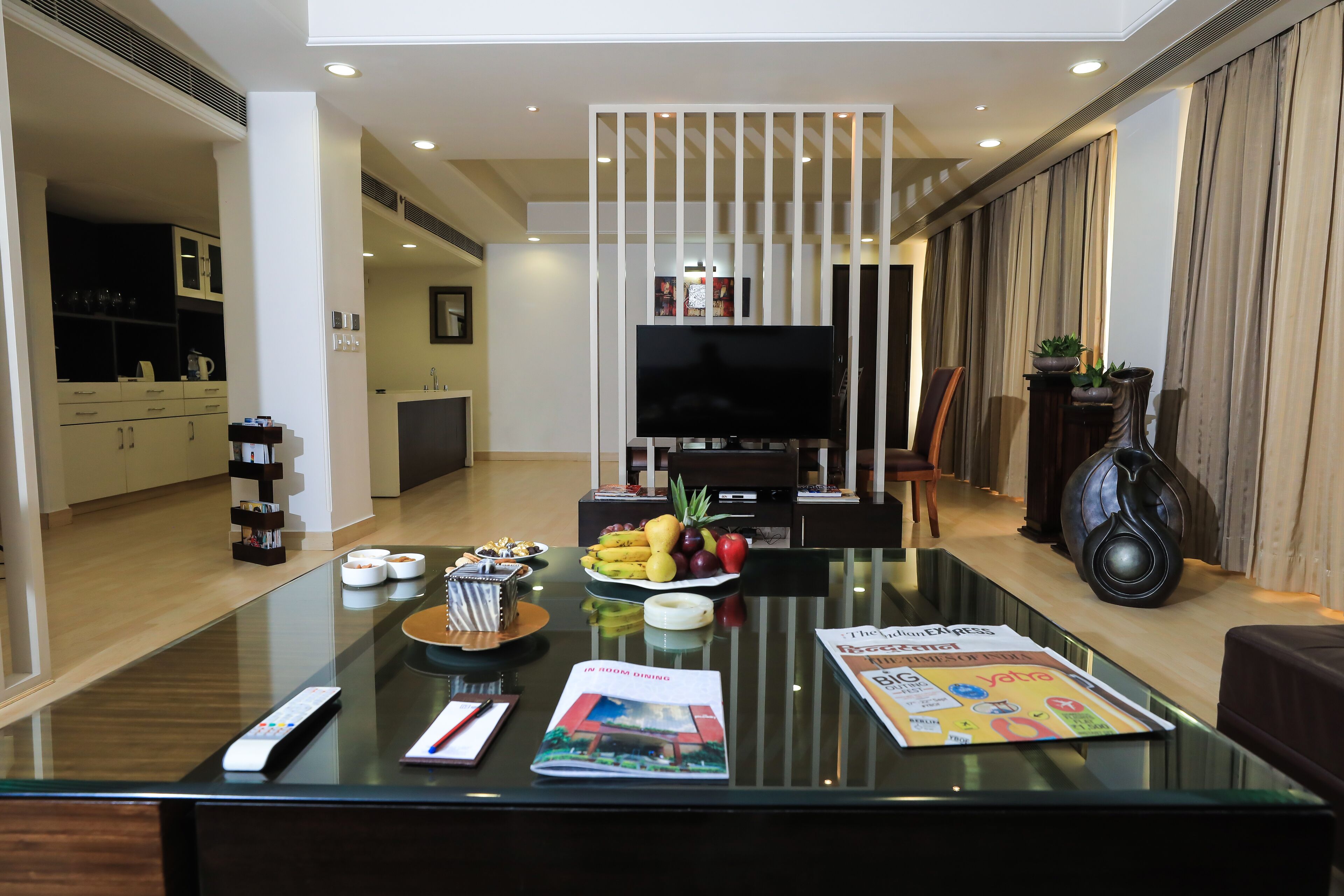 A modern living room with a glass coffee table, a flat-screen TV, and a dining area with a fruit bowl.