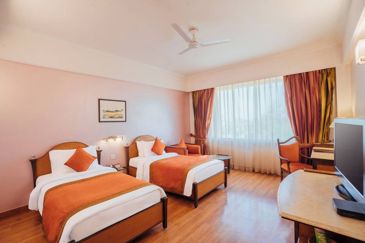 Quarto de hotel com duas camas, uma escrivaninha, televisão e ventoinha no teto.