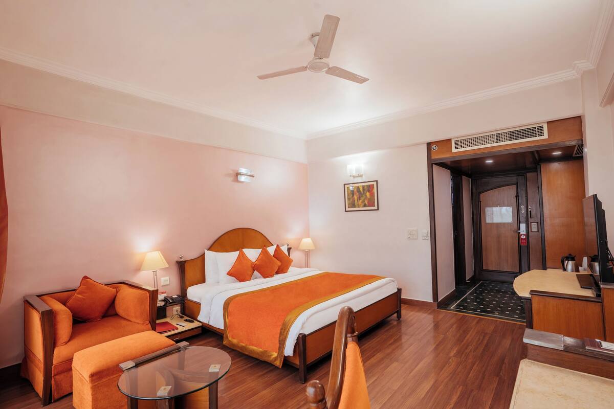 Quarto de hotel com cama, sofá, mesa pequena, ventoinha no teto e elevador.