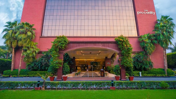 A hotel with a red brick facade, a large glass entrance, and palm trees in the foreground. - The Piccadily Lucknow (Lucknow)