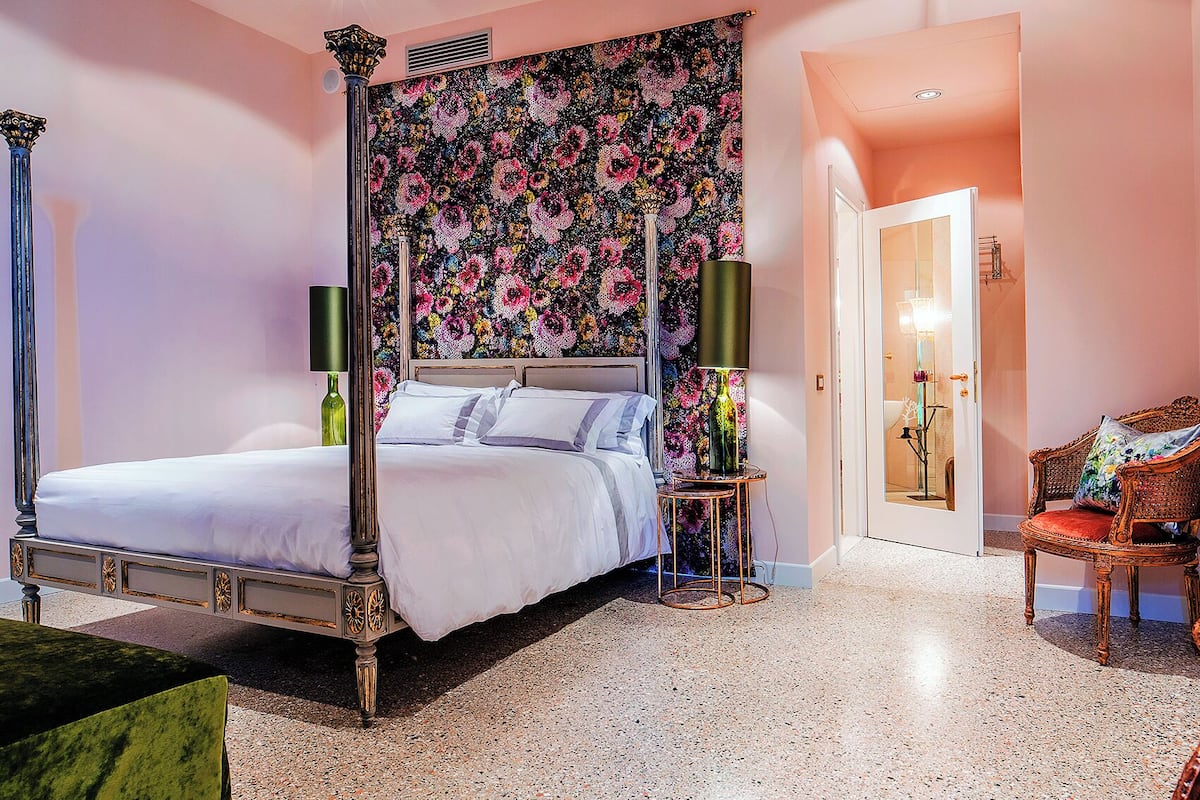 A four-poster bed with a floral headboard, a side table with a lamp, a wicker chair, and a door leading to another room.