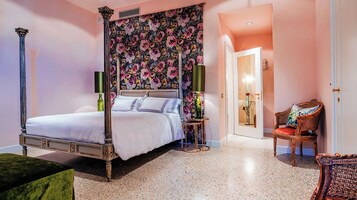 Un letto a baldacchino con testata fiorita, un comodino con lampada, una poltrona in vimini e una porta che dà su un'altra stanza.