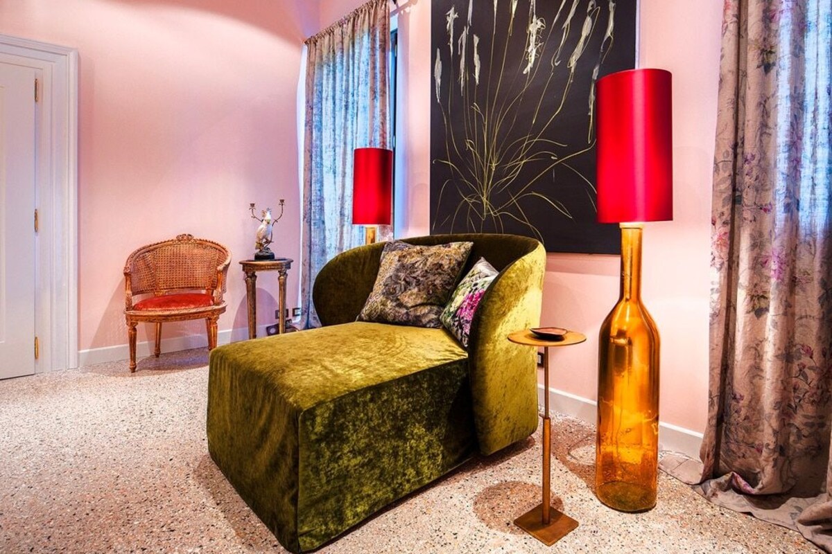 A room with a green velvet sofa, a red floor lamp, a tall glass bottle, and a large abstract painting on the wall.