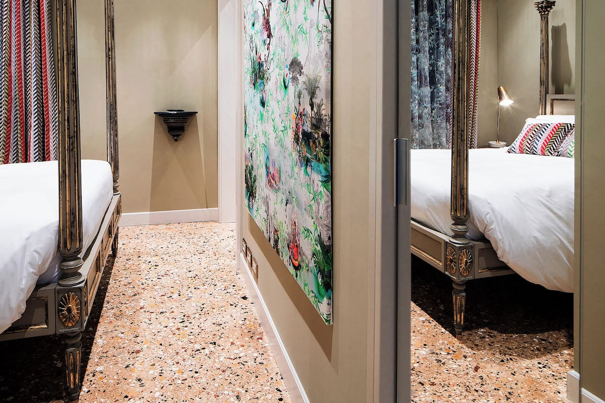 A hotel room with a large mirror, a bed with a headboard, a wall-mounted artwork, and a floor with a speckled pattern.