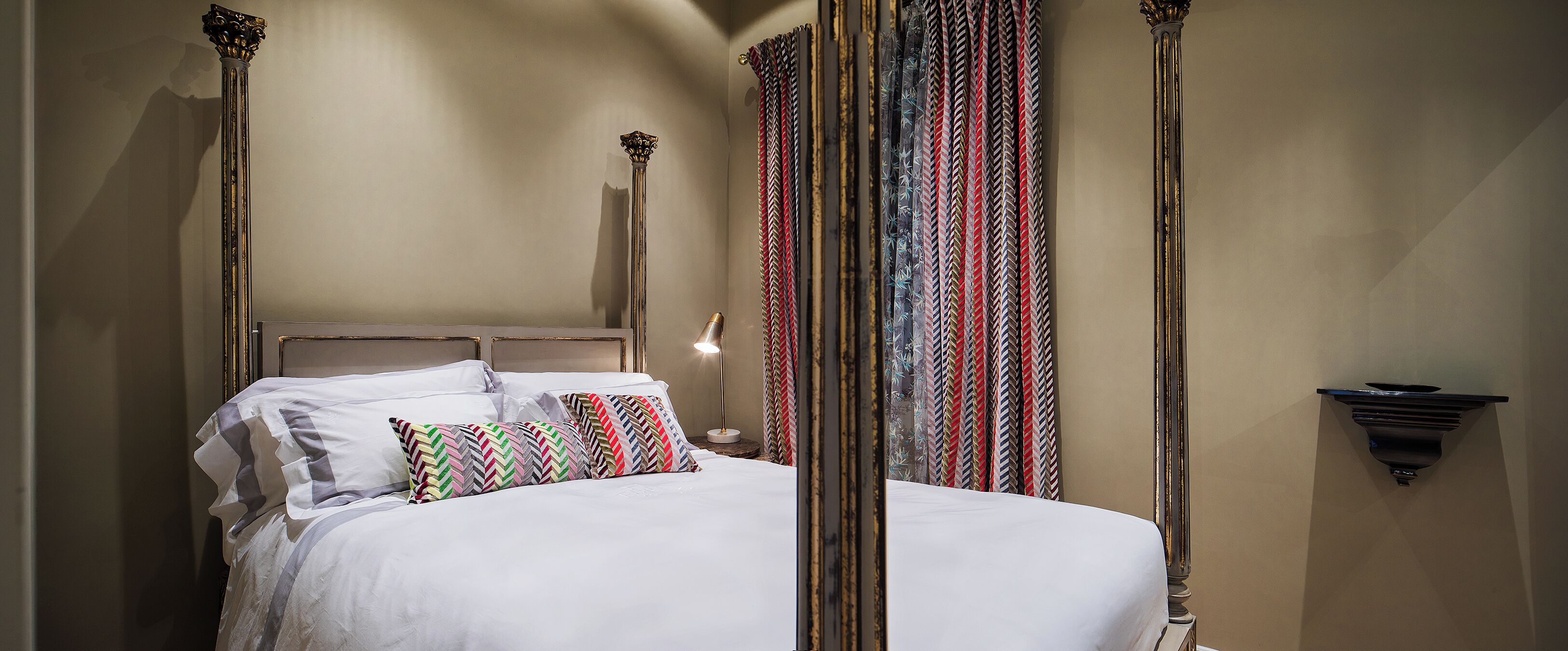 A neatly made bed with a headboard, a bedside lamp, and patterned curtains.